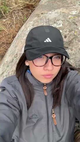 The video shows a woman lying down on a rock in a mountainous area. The woman is wearing a grey jacket, a black baseball cap with the Adidas logo, and glasses. She is lying on her back with her eyes closed. The background shows a wide valley with rolling hills and mountains in the distance. The sky is cloudy. The camera pans to show the landscape around the woman, revealing dry grass and bushes. In the final shot, the camera pans to show another person sitting on a rock in the distance.