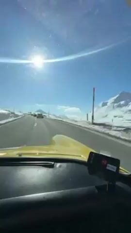 A yellow car travels down a highway with a bright blue sky and snowy mountains on either side. The sun shines brightly, reflecting off the snow. The camera is inside the car and shows the dashboard, a small electronic device, and a sticker on the windshield. The view through the passenger-side window shows snow-covered mountains and roadside markers.