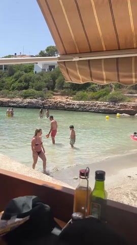 A group of people are wading in clear, shallow water at a beach. The water is calm and the bottom appears sandy or rocky. In the foreground, a person in a bikini is walking out of the water towards the beach. Behind them, a man in red shorts and a boy in green shorts are walking further into the water. Further back in the water, other people are visible, some appearing to be swimming or playing. The beach has a rocky shoreline and is bordered by green, overgrown hills. There is a building with white walls and a dark roof visible on the hillside. Overhead, a brown awning provides shade. In the extreme foreground, on a wooden surface, are two bottles, one clear with a red cap and another green with a black cap.