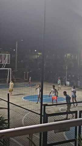 Here is a detailed description of the video.

The scene unfolds on an outdoor basketball court at night, lit by overhead lights. The court is enclosed by a chain-link fence. A volleyball net is stretched across the middle. On the court, about eight people are playing volleyball, some casually dressed in shorts and t-shirts. The court is painted with a blue circle in the center.

A basketball hoop stands at one end of the court, with a makeshift backboard. Behind the court, there is a row of parked scooters or mopeds, and some onlookers are visible.

The camera is positioned from an elevated perspective, likely from a balcony overlooking the court. A railing is visible in the foreground. The video is somewhat grainy and the lighting is not bright, but the action is clear.

The overall atmosphere is relaxed and casual, suggesting a friendly game among acquaintances or community members.
