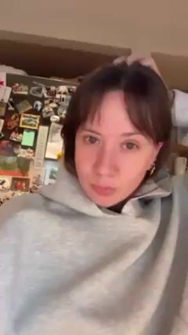 A woman in a gray hoodie is facing the camera. She has short brown hair with bangs and earrings. She winks, moves her bangs, and makes a kissing face. The background includes a refrigerator covered in magnets and other kitchen storage.