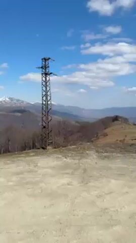 The video opens on a scene of a slightly elevated, unpaved, sandy or gravelly ground with a view overlooking a wide, mountainous terrain under a partly cloudy, bright blue sky. In the immediate view, a tall, metal, lattice-like power line structure stands to the left, with the terrain sloping down to reveal distant mountains and a valley below. The camera then pans to the right to reveal a truck parked at the edge of the scene. It’s loaded with a large number of bicycles securely attached to a rack mounted on the back of the truck. The truck appears stationary, with its presence casting a long shadow on the ground. The video is shot in daylight and suggests an outdoor recreational setting.