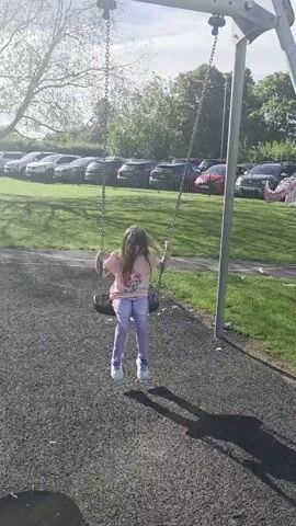 A young girl with long brown hair is swinging on a swing set in a park. She is wearing a pink t-shirt with a graphic on it and purple tie-dye pants with white sneakers. The swing set is made of metal with grey chains. The ground around the swing set is covered in dark grey mulch. In the background, there is a grassy area with trees and a parking lot with several cars. The lighting suggests it is daytime, possibly late afternoon, with the sun casting shadows. The girl is in motion, swinging back and forth.