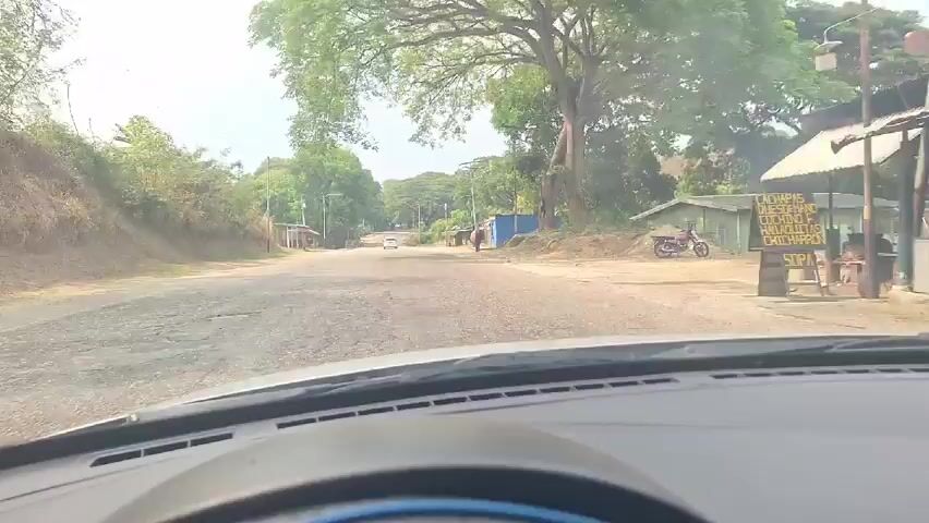 The video shows a view from inside a car, looking out onto a somewhat rural road. The road is paved, but the shoulder appears to be dirt and gravel. There are houses and small buildings on either side of the road, and lush green trees line the roadside. A person stands under the awning of a blue building on the right side of the road. Another car, a white SUV, passes the camera vehicle. The sky is overcast, suggesting it's either early morning or a cloudy day. The camera is static, providing a driver's perspective.