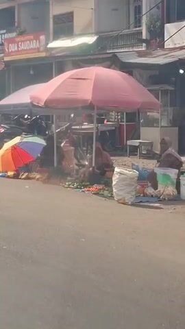 The video shows a street scene with several vendors selling produce under umbrellas. In the foreground, a black Mitsubishi Xpander vehicle is driving by. The street is paved and appears to be in a commercial area with shops and storefronts lining the road. There are also motorcycles parked along the side. The vendors are sitting on the ground next to their goods, which include fruits and vegetables. The overall atmosphere is that of a local market.