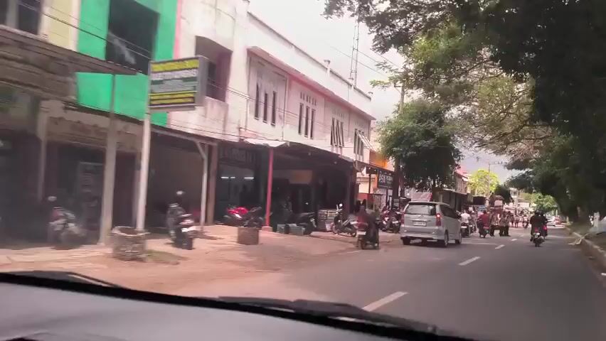 The video shows a street scene in Indonesia. From the perspective of a car driving down the road, people dressed in formal uniforms, some wearing headscarves, are gathered on the side of the road in front of buildings. Many motorcycles are parked nearby. Other cars and motorcycles are also present on the street. Trees line the side of the road. The buildings appear to be two or three stories tall and have awnings in front of some of the storefronts. The sky is overcast.