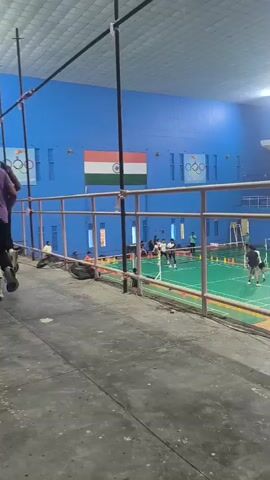 This is a video taken from a vantage point above an indoor sports facility. The walls of the facility are painted a vibrant blue. Prominently displayed on one wall is the Indian flag, with the Olympic rings logo visible nearby. The floor of the sports area is a patchwork of green, pink, and blue mats, suggesting it's a multi-purpose facility for sports like badminton and martial arts.

Several individuals are engaged in activities on the mats below. In the background, what appears to be a badminton court is set up with a net. There are people playing badminton, some individuals are practicing, and others appear to be observing. Further into the facility, the blue and pink mats indicate areas for other sports, possibly taekwondo or karate, with people practicing various stances and movements. The lighting in the facility is bright, suggesting it is daytime or the facility is well-lit for evening use.

A metal railing runs along the lower part of the frame, obscuring some of the view. The foreground is a concrete surface. The overall impression is of a busy, active sports complex. The presence of the Indian flag and Olympic rings suggests a connection to national or international sporting events, or perhaps a training facility for athletes. The mood appears to be one of focused activity and sportsmanship. There is visible text on some of the sporting equipment, including "DONIC" on blue boards along the edge of one court.