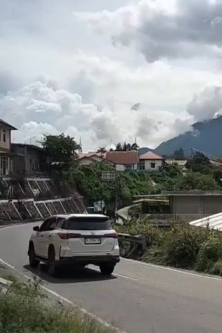 The video shows a slightly elevated, slightly angled view of a paved road winding through a hillside community. In the foreground on the right, a portion of a dark-colored vehicle is visible. On the left side of the road, a white SUV is driving away from the camera. Further down the road, a scooter with two passengers is also driving away. The buildings in the community are varied in style and color, with some appearing to be houses and others possibly institutional buildings. Terraced retaining walls are visible on the left side of the road. Vegetation, including trees and shrubs, is abundant throughout the scene. The background features a mountainous landscape partially obscured by dramatic, billowing clouds. The sky is overcast. The overall impression is of a scenic, semi-rural environment with active transportation.