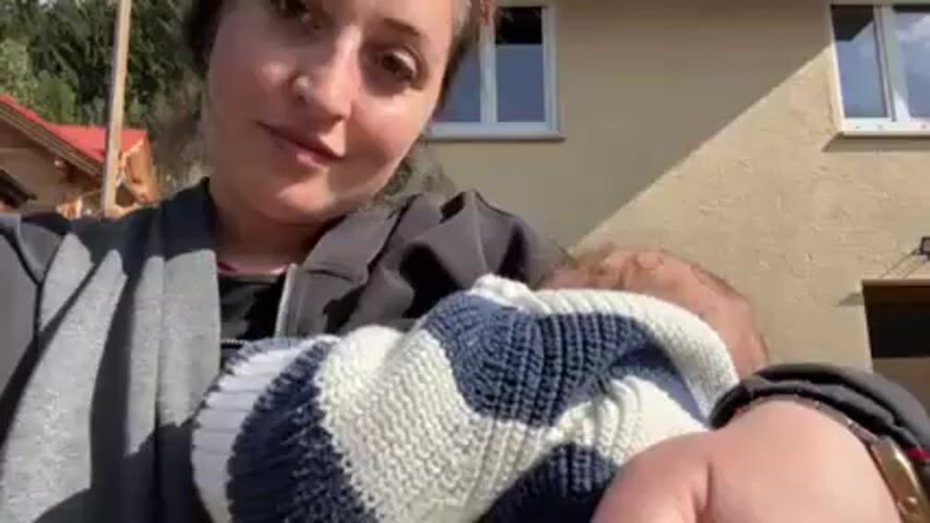 The video shows a woman holding a baby, moving it out of view and looking directly at the camera. The woman is wearing a black jacket with a gray scarf, and a red necklace. She has brown hair pulled back and is standing outside in front of a building with light brown siding. There is a window with a closed shutter, and some vegetation in the background. In the first frame, the baby is wrapped in a blue and white striped sweater. The woman smiles slightly and makes a pouting face toward the camera.