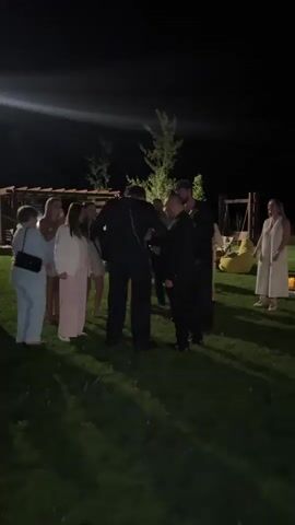 A group of people are gathered outdoors at night. The grass is illuminated by lights. In the foreground, a person in a black outfit, possibly a costume, stands facing away from the camera, talking to another person in a black suit who is facing towards the camera. Several other individuals, appearing to be women in dresses and a person in a light-colored outfit, are standing to the left of the frame, observing the interaction. In the background, there are structures that resemble a wooden pergola or gazebo, and some yellow objects that could be candles or lights scattered on the grass. A large, blurry light source is visible in the upper left corner of the frame. The overall atmosphere suggests an outdoor event or gathering at night.