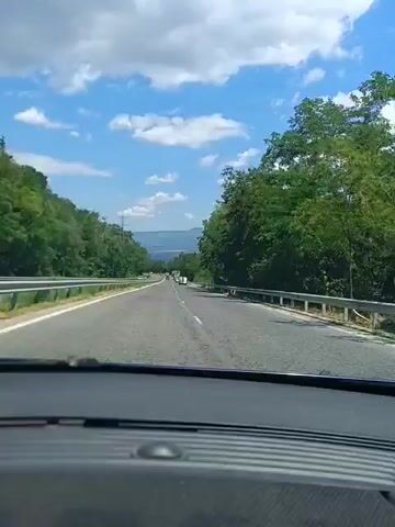 The video captures a first-person view from inside a vehicle, driving on a highway. The road is paved, with two lanes visible in the direction of travel and a divided lane for oncoming traffic. Guardrails line both sides of the highway, separating the road from lush green trees and vegetation. The sky is blue with scattered white clouds, suggesting fair weather. In the distance, mountains or hills are visible, adding to the scenic landscape. Several other vehicles, including cars and trucks, can be seen on the highway, indicating moderate traffic. The dashboard of the vehicle is visible in the foreground. The overall impression is of a leisurely drive through a natural, wooded environment.