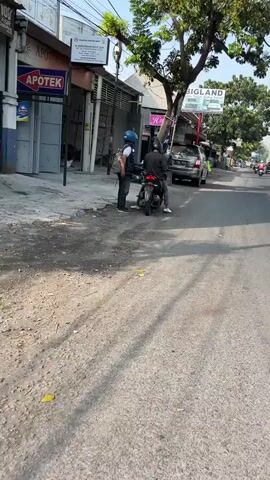 The video shows an outdoor scene on a paved road with a sidewalk. Two individuals are present. One person, wearing a blue helmet and a backpack, stands on the sidewalk next to a parked motorcycle. The other person, wearing a black helmet and black clothing, is seated on the motorcycle. The motorcycle is facing the camera. In the background, there is an apothecary with signage, trees, and a building with a sign that reads "BIGLAND". The lighting suggests it is daytime.