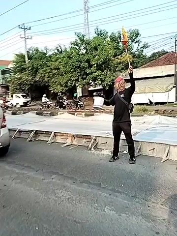 The video shows a road construction site with various vehicles passing by. At the start, a man in a black outfit and a red cap is seen directing traffic with a flag. The camera then focuses on a silver car passing by, followed by a yellow truck. The scene shifts to a wider view of the street, showing a gas station, various vehicles including motorcycles, and utility poles with power lines. Overall, the video provides a glimpse of a typical street scene in an urban area undergoing road construction.