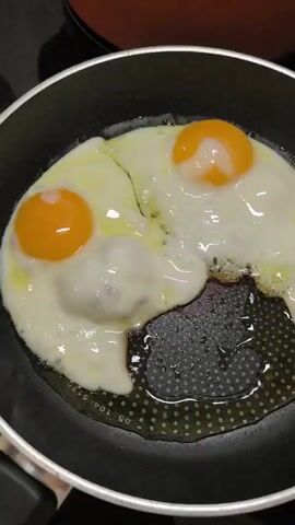 A pair of fried eggs with bright orange yolks are in a black non-stick frying pan. A slice of red tomato is placed between the two eggs. A hand with light skin and short fingernails is reaching in to adjust the tomato. The eggs are being cooked in oil, which is visible in the pan. The background is dark and out of focus.