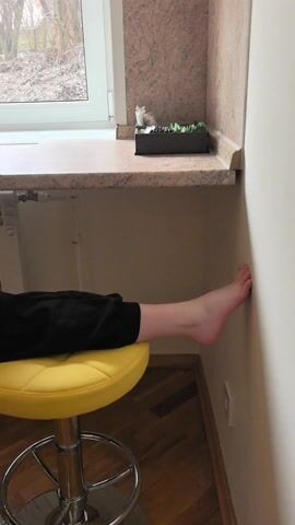 The video shows a young girl sitting on two yellow bar stools in a split position at a kitchen counter. She is facing away from the camera, looking out of a window at trees without leaves. She has long brown hair braided down her back. She is wearing a long-sleeved black top and black pants. The counter has a microwave and kettle on it. The room appears to be indoors, possibly in a house or apartment.