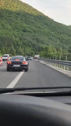 The video shows a view from inside a vehicle, looking forward at a highway. The road is a multi-lane highway with a shoulder bordered by a metal guardrail. On the left side of the road, there are traffic cones and a white van. Ahead, a black Porsche Cayman is driving in the right lane, followed by a grey hatchback. In the distance, a large white truck is visible. The highway is surrounded by a lush green, tree-covered mountainside. The sky is a pale blue with some clouds. The car is moving slowly, suggesting traffic or road work.