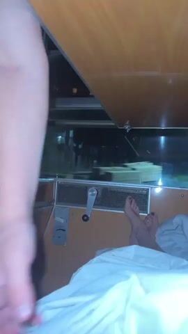 A first-person perspective video shows a person's arm and hand in the foreground, partially obscuring the view of the background. The background appears to be inside a train compartment. A window shows a blurred nighttime view of lights from a city or industrial area. In the bed on the right side of the frame, a person's bare feet are visible, with dark nail polish on their toes. There is white bedding covering most of the bed. A blue water bottle and a red bag are on a shelf above the window. The overall lighting is dim.
