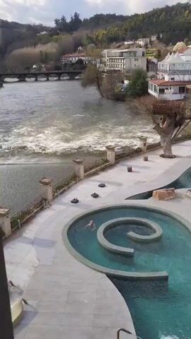 The camera pans around to show a resort swimming pool next to a fast-moving river. The pool is made up of several sections, including a spiral-shaped pool and a jacuzzi-like pool. There is a person swimming in the spiral-shaped pool and another person soaking in the jacuzzi-like pool. The resort buildings are visible in the background.