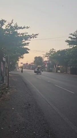 A person is seen walking along the side of a paved road. Several motorcycles are present on the road, with one rider wearing a red helmet. Trees line the sides of the road, and there are buildings and signs in the background. The sky is a hazy yellow, suggesting either sunrise or sunset.