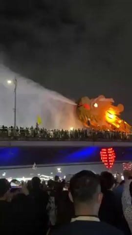 At night, a large, illuminated golden dragon head sculpture on a bridge is shown exhaling a stream of water, seemingly in a show. People are standing on the bridge and on a lower level, watching and recording with their phones. The bridge is decorated with red lanterns, and the sky is dark. Small Japanese flags are visible among the crowd.