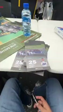 The video shows a crowd of people standing around a table covered with postcards of a race car driver, Ariel Levi. Some people are signing autographs on the postcards and other items that people hold out. A poster advertising a race event at the Sachsenring in Germany is visible. Some people are wearing jackets and hats, suggesting that the weather is cool or rainy.