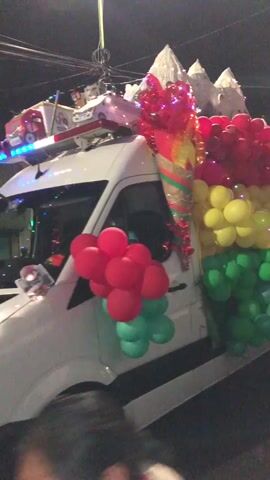 The video shows a white van decorated for a parade or celebration. The van is covered in balloons arranged in horizontal stripes of red, yellow, and green, resembling a flag. In the front of the van, a large bouquet of red balloons is attached to the driver's side door, with a smaller cluster of red and green balloons below it. The van is decorated with a centerpiece on top that appears to be a large, festive structure with white, snow-capped mountain shapes. Small lights are interspersed among the balloons. In the background, there is a banner with Spanish text that reads "SECRETARIA MUNICIPAL DE SALUD INTEGRAL Y CIUDAD INTELIGENTE". The scene appears to be at night or in low light conditions, with streetlights visible in the background.