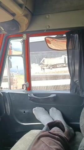 The video shows a view from inside a truck cab, looking out the window. In the foreground, a person's legs are visible, clad in grey socks and brown pants, resting on the seat. The person's feet are crossed. Outside the window, a construction scene is visible. A white dump truck with the text "UZTBM" on its side is parked. Several people are working near the truck, laying down paving stones. One person is standing by the truck's rear tire, and two others are in front of the truck, seemingly placing stones. The ground is covered with gravel and more paving stones. In the background, there is a large dark structure, possibly a building or a wall, and some greenery on a hill. The sky is not visible.