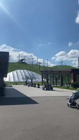 The video shows an outdoor scene on a sunny day with a bright blue sky and scattered white clouds. In the foreground, there is an asphalt pathway with some manhole covers visible. To the left, a building with a wooden overhang and a modern stone facade is seen. To the right, further down the path, there are more modern buildings with glass windows and stone exteriors.

In the middle ground, a large, white, tent-like structure is set up on a grassy slope. Behind the tent, a ski lift with chairs going up the hill is visible, suggesting a ski resort even in warmer weather, possibly for artificial snow or a summer attraction. There are trees on the slope and in the background.

Several people are scattered throughout the scene. Some are walking on the pathway, others appear to be sitting on benches near the tent. A few individuals are on bicycles or skateboards. A white van with a green logo is parked near one of the buildings. There is also a small blue and pink multi-seat cart with wheels near the building on the left. The overall impression is a public recreational area or resort.