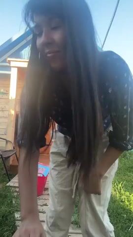 A woman with long dark hair, wearing a dark blue patterned shirt and light-colored pants, is standing on a wooden path in front of a wooden house with a blue roof. She is bending down and picking up a red bucket. Inside the bucket are two items, one appears to be a book with a pink cover and the other is a light blue object. She then walks up the steps onto the porch of the house and places the red bucket on the porch. She appears to be in an outdoor, rural setting with green grass and trees visible in the background. The lighting suggests it is daytime.