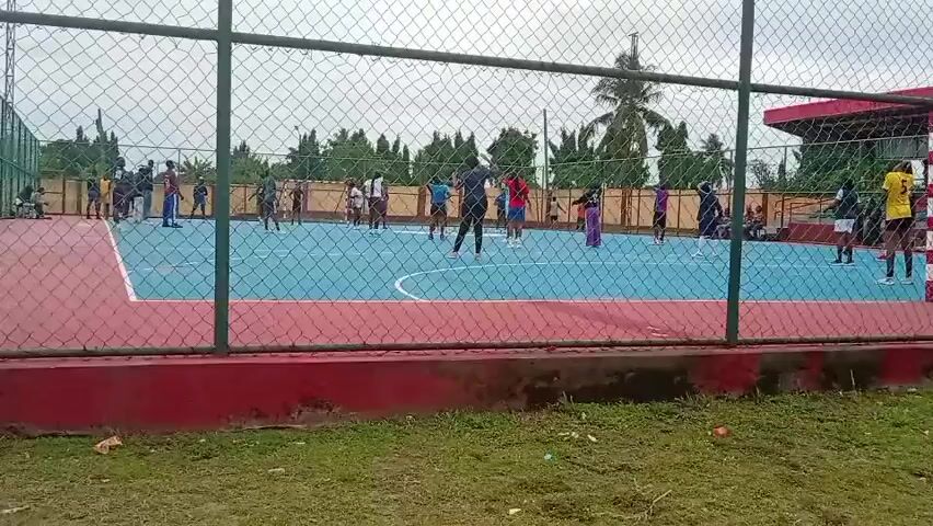The video depicts a group of people on an outdoor handball court, surrounded by a chain-link fence. The court's playing surface is primarily blue, with red sections at either end and white lines marking boundaries. Players are scattered across the court, engaged in what appears to be a practice or training session. They are wearing various colored shirts, including white, blue, and yellow.

The environment surrounding the court includes a green grassy area in the foreground, with trees and other foliage visible in the background. A building with a red roof can be seen behind the fence. The sky overhead is overcast, suggesting a cloudy day.