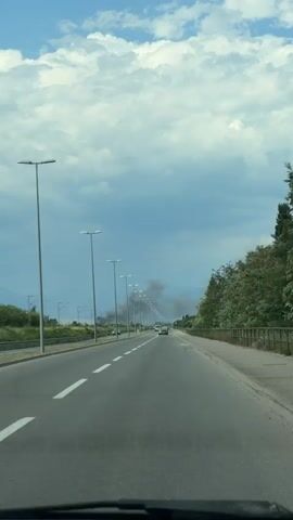 The video shows a POV shot of a person driving on a highway with a view of smoke rising in the distance. The smoke appears to be coming from a fire, as it is dark and billowy, and obscures the horizon in the background. The highway is lined with streetlights on the left side of the road, and there is a small number of cars on the road. To the right of the highway, there is a sidewalk and a fence, followed by trees and vegetation. The sky is cloudy, with some patches of blue visible. The time of day appears to be afternoon, based on the lighting. The overall mood of the video is somewhat ominous due to the presence of the smoke, suggesting an emergency or a significant event is unfolding. There is no visible text in the video.