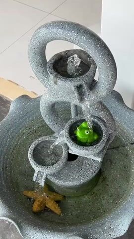 The video shows a close-up, low-angle shot of a small, tiered indoor fountain made of speckled gray material. Water flows from the top tier, cascading down to a middle tier where a bright green, cartoonish frog figurine sits in a circular basin. The water then flows from the middle tier to a lower tier, and finally spills into the main basin at the bottom. A small, tan, starfish-shaped object is submerged in the water in the main basin, near the lowest spout. The water is clear, and the movement of the water is the primary focus. The background is out of focus and appears to be an indoor setting with light-colored walls and flooring. The lighting is bright, suggesting an indoor space with artificial or natural light. There are no people or animals other than the figurine visible in the video.