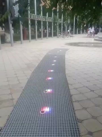 This is a video of a public walkway in Atyrau, Kazakhstan. The walkway is paved with interlocking concrete stones and features a long, raised metal grate running down its center. Embedded within the grate are several circular lights that emit colored light, creating a visually interesting effect. The lights appear to be cycling through different colors, predominantly red, pink, blue, and white. In the background, there are trees, some light poles, and other people walking. Two bicycles are leaning against a railing in the middle ground. Further in the background, a fountain is visible with water spraying upwards. The overall scene suggests a pleasant daytime setting, likely in a park or a pedestrian area. The lighting of the walkway indicates it might be designed for evening or night use as well. There is text visible on a sign in the background which appears to be in Cyrillic script.