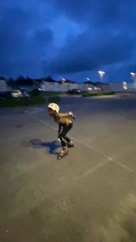 A person wearing rollerblades and protective gear is rollerblading in a parking lot. The person is wearing a helmet, knee pads, and elbow pads. They are wearing a dark outfit with white and orange accents. The rollerblades are mostly black. The parking lot is paved and has white lines painted on it. There are buildings and cars in the background. The sky is dark blue. The person is seen rollerblading at different angles.