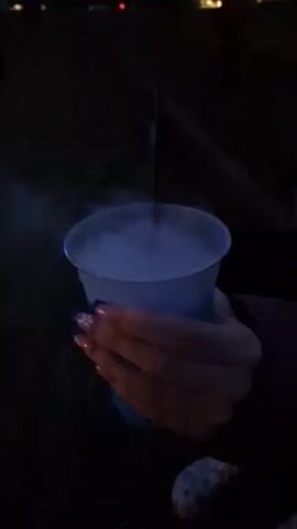 A close-up shot of a hand holding a translucent blue cup filled with a light blue liquid, from which a plume of white smoke or vapor is rising, obscuring the top of a dark straw. The background is dark and out of focus, suggesting an outdoor setting at night, possibly a yard or patio. The hand has long fingernails painted with a dark, possibly blue or purple, polish with glitter accents. The cup has a textured blue sleeve around its lower portion.