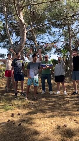 Six men of various heights are standing in a clearing between trees, with pine cones scattered on the ground. The man in the center is holding a phone in front of him, facing the camera. All the men are wearing casual clothing. The man on the left is in a beige tank top and red shorts. Next to him is a man in a black graphic t-shirt and navy shorts. The man in the center has a gray shirt with a faded white portion at the bottom and blue patterned shorts. To his right is a man in a green striped shirt and jeans. Another man is to his right wearing a gray hooded sweatshirt and black shorts. The man on the far right is in a black shirt and navy shorts. The men are all moving their arms in a seemingly coordinated dance. There are trees behind and above them, and decorations tied around the trees. The sky is visible through the trees.