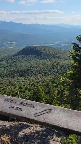 The video shows a scenic mountain vista from a wooden lookout post. In the foreground, a wooden railing has signs attached to it. One sign reads "MONT ROYAL 94 Km" with an arrow pointing to the right. Another sign reads "LAC CHAMPLAIN 57 Km" with an arrow pointing to the right. The background consists of rolling green hills covered in dense forest, with distant mountains under a partly cloudy blue sky. The camera pans slightly, offering a wider view of the landscape.
