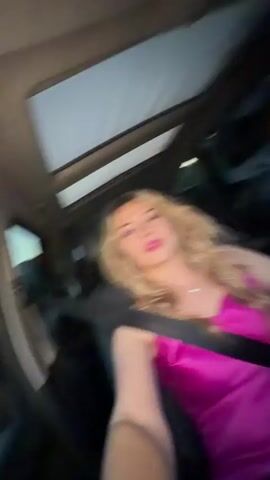 A woman with curly blonde hair, wearing a hot pink tank dress and a seatbelt, is sitting in the front passenger seat of a car. She is looking at the camera and singing. A man is partially visible in the driver's seat. The car has a sunroof.