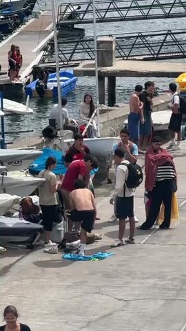 The scene takes place at a marina or harbor on a sunny day. There are multiple small sailboats docked, along with other various watercraft. A group of approximately a dozen young people are standing on a concrete pier. Some of them are changing clothes and toweling off. They are mostly dressed in casual clothes. There is a flag of Madeira flying and buildings in the distance. A couple more people walk by in the foreground.