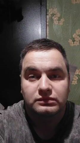 The video is a selfie taken by a man indoors. The man is in the foreground, looking at the camera. Behind him, on the left, is a dark object, possibly a closet or a piece of furniture. On the right, a wall is visible with green floral wallpaper. The man is wearing a grey t-shirt. In the background, to the left of the man, a ginger cat is sleeping on a bed. The man blinks and gestures with his finger.
