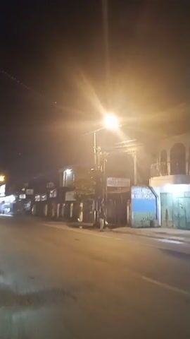 The video shows a nighttime street scene, likely in a somewhat rural or less developed area, with a single bright streetlight illuminating the foreground. A paved road runs horizontally across the bottom of the frame. On the left side of the road, there are several buildings with signs, appearing to be shops or businesses. Some of these signs are in a language that is not English. On the right side of the road, there are more buildings, including one with a balcony. A person in a light-colored shirt and dark pants is visible walking on the sidewalk. Further down the road, a large truck with its headlights on is moving from left to right. The overall impression is one of a quiet, perhaps dimly lit commercial street at night. The footage is somewhat shaky, suggesting it was filmed handheld while moving.