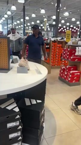 The video shows a shoe store with several people browsing. In the foreground, a light-colored shoe is displayed on a round table with black Adidas boxes underneath. Several people are walking in the background, some of whom appear to be looking at shoes. The store is well-lit with fluorescent lighting and has shelves stocked with shoe boxes. A "Back to School" sign is visible in the background. The overall atmosphere appears to be a typical retail environment.