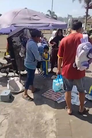 A group of people are gathered around a market stall on a sunny day. A man wearing a red shirt and a purple backpack is standing with his back to the camera, facing a young girl in a blue and yellow outfit. To the left, another man in a denim jacket is standing next to the stall, which is covered by a purple awning and displays various items. Two young children in blue and yellow outfits are also near the stall. In the background, other people and a motorcycle are visible, suggesting a busy outdoor marketplace.