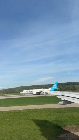 The video shows an aircraft on a runway from the vantage point of another aircraft. The view shows the wing of the aircraft the camera is in. On the runway ahead, another aircraft with an "AirJet" logo is visible. In the background is a green, hilly landscape under a blue sky with scattered clouds. The runway is surrounded by green grass. The video appears to be taken during daylight hours. The other aircraft is moving.