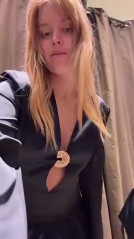 A woman with long blonde hair stands in a dressing room. She is wearing a long-sleeved, black, satin-like dress with a cutout at the waist and a plunging neckline. The dress has a large, round, gold-colored clasp at the neckline. She turns slightly to show the side cut-out. The dressing room has a beige curtain background. Another person is partially visible in the mirror.