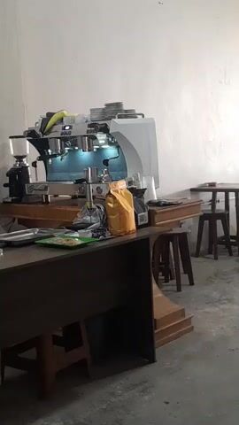 The video shows an espresso machine and a coffee grinder on a counter. There are also bags of coffee beans, cups, and other coffee-making equipment. A dark wooden table and stools are visible in the foreground. The scene appears to be a coffee shop or a home barista setup. The lighting is bright, suggesting it is daytime or well-lit indoors. The wall behind the counter is white.
