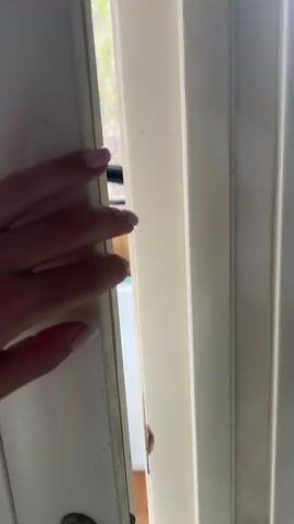 A person's hand with light-colored nail polish opens a white door, revealing a cluttered enclosed balcony or sunroom. The initial view shows a white car door frame and a white car door leaning against a wall, next to stacked cardboard boxes, plastic bags, and a metal tin. One of the boxes has "AKKA" visible on its side. A small white stool is also present. Through the large windows, tall residential buildings and trees with pink blossoms are visible, suggesting a springtime urban or suburban setting under an overcast sky. The camera then pans right, showcasing the balcony's wooden-paneled walls. Under the white stool, what appear to be weights or books are stored. The pan concludes on an orange-colored cat, likely a Scottish Fold, sitting calmly on a light brown blanket draped over a wooden chest or bench. The cat looks directly at the camera and meows.