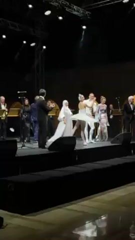 A video shows a stage performance. The set consists of a series of raised black platforms. On the stage, several formally dressed individuals stand behind a ballet dancer and her partner in white tutus and tights, who are holding hands. A man in a suit claps his hands. Men with instruments stand to the left of the stage, while women in long gowns stand to the right. Stage lights are visible overhead, and the background is dark. The performance appears to be taking place indoors. The audience is visible in the foreground of the video.