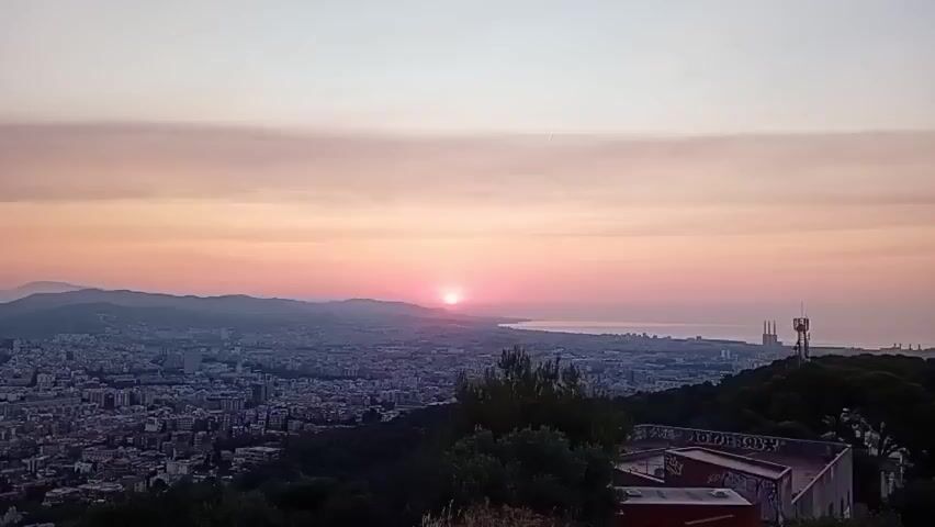 The video shows a panoramic view of a city and coastline during a sunrise. The sun, a bright pink orb, is low on the horizon, casting a soft pink and orange glow across the sky and reflecting on the water. The city stretches out below, with buildings densely packed together, and hills visible in the distance. There are some structures and antennas visible on a hilltop in the foreground, as well as trees. The overall impression is one of a serene and expansive dawn over an urban landscape.