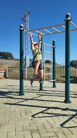 A person wearing a green tank top, denim shorts, and a straw hat is hanging from a monkey bar. They are in mid-motion, with one leg bent and the other extended. The monkey bar is part of an outdoor exercise equipment set, featuring green metal poles and a silver horizontal bar. The ground is paved with interlocking concrete tiles, and shadows are cast from the equipment. In the background, a green metal fence runs along a grassy embankment. Further back, a tall, red and white pylon structure with wires is visible against a clear blue sky. The scene appears to be an outdoor park or recreation area on a sunny day.
